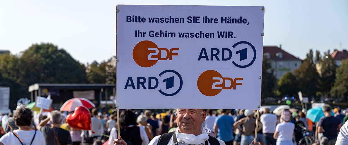 Ein Demonstrant gegen Corona-Schutzmaßnahmen hält ein Schild hoch, auf dem die Logos von ARD und ZDF zu sehen sind mit der Aufschrift "Bitte waschen Sie Ihre Hände. Ihr Gehirn waschen wir"
